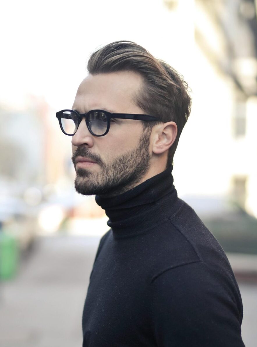 A fashionable man in a black top and glasses poses in an urban street with parked cars.